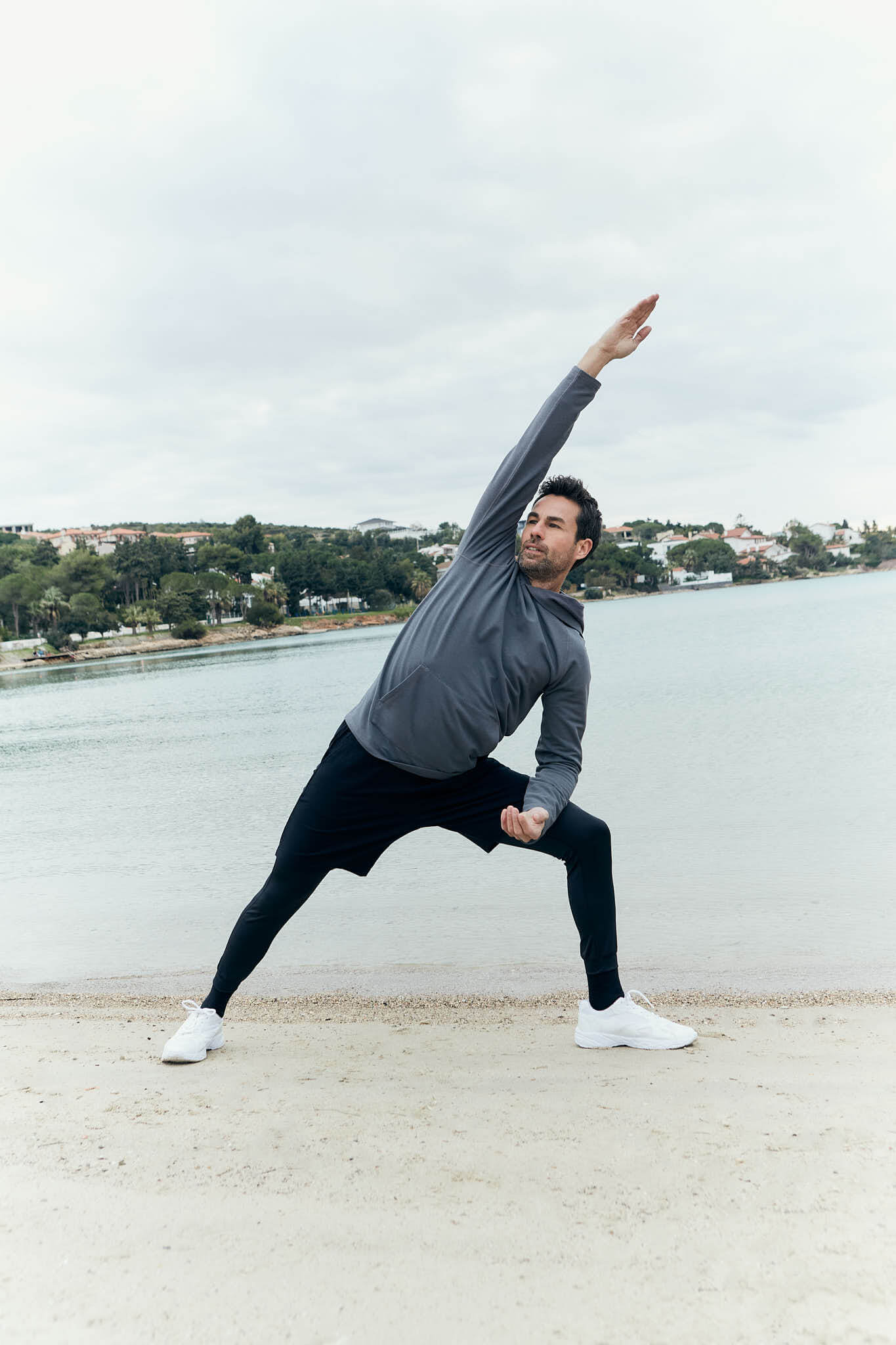 Man in a gray hoodie and black athletic leggings performing a side stretch on a sandy beach, with a calm body of water and greenery in the background under an overcast sky.